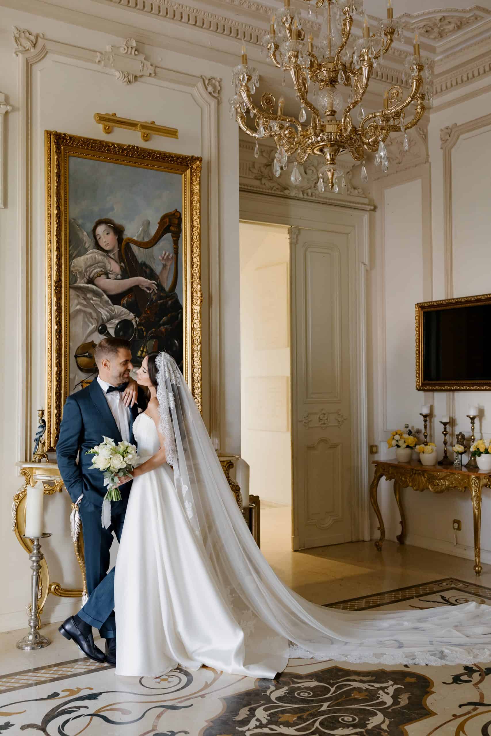 Elegant wedding couple in ornate historic venue with classic decor and grand chandelier.