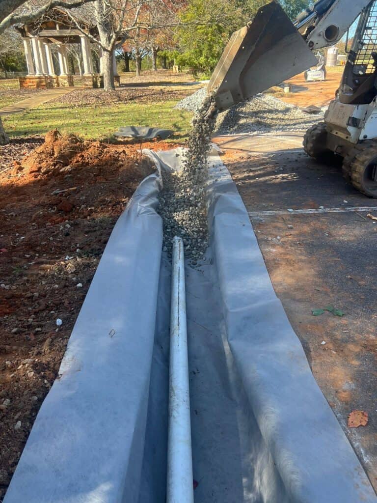 Crushed stone being poured from a skid steer over a 4-inch perforated pipe in a geotextile-lined French drain trench at the UGA GSI demo site