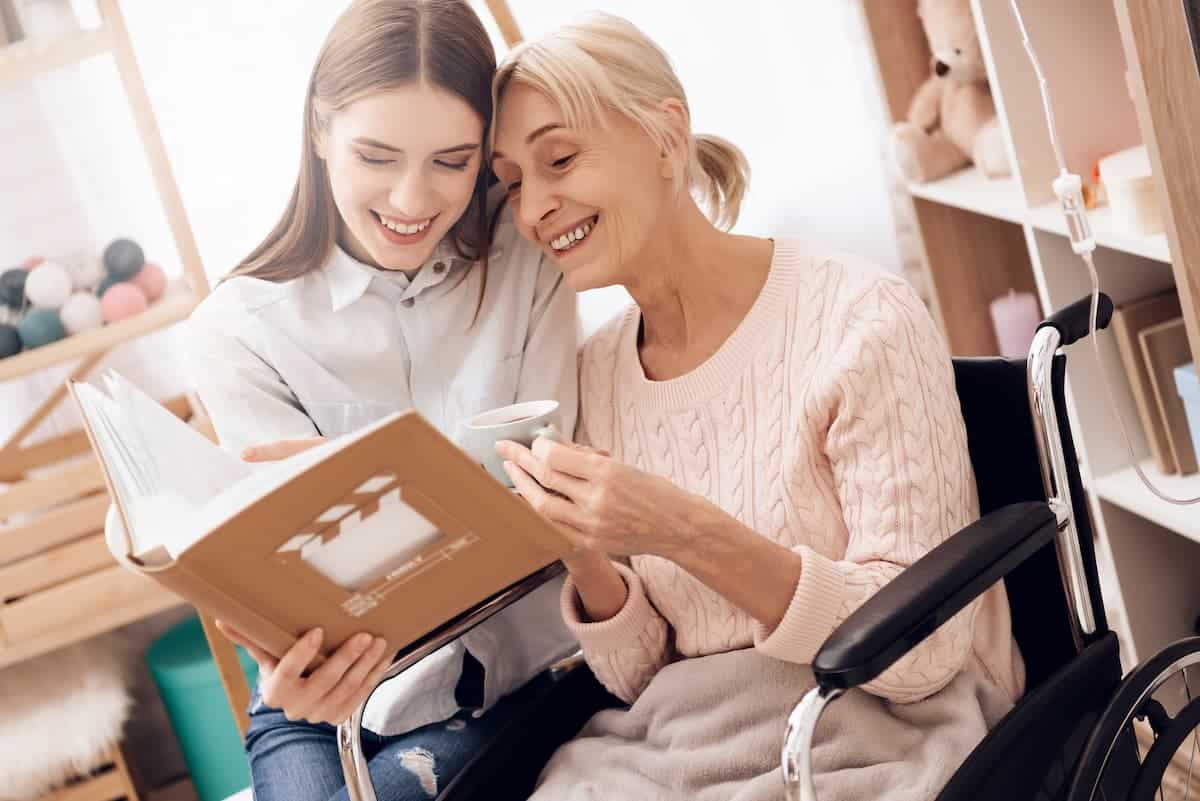A young woman reading a book with an elderly woman in a wheelchair while they both smile