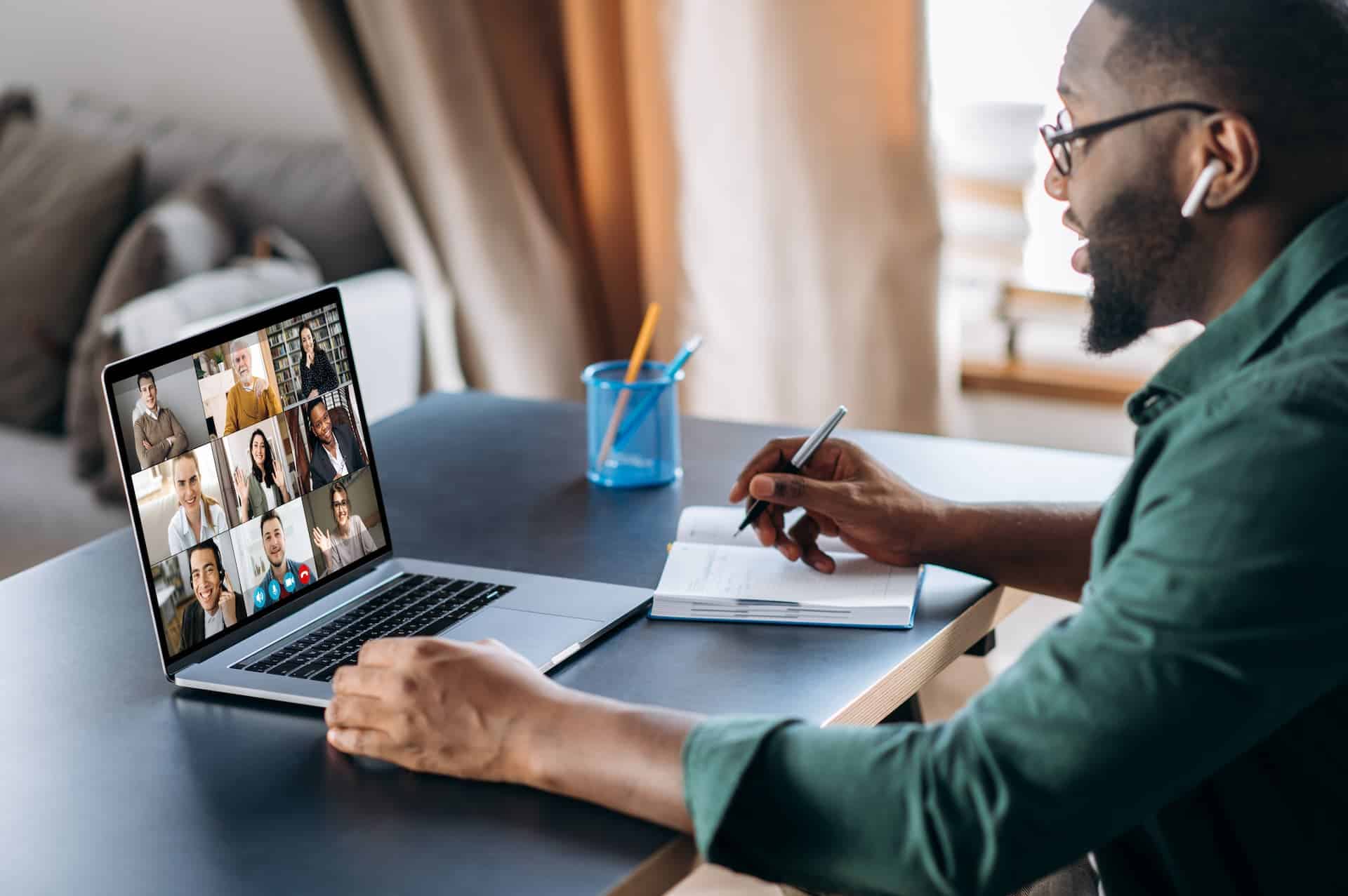 A man on a group video call webinar on his laptop while taking notes on some paper