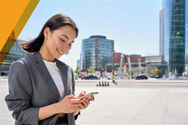 A woman in a gray jacket looking at her phone and smiling in a business plaza on a sunny day.