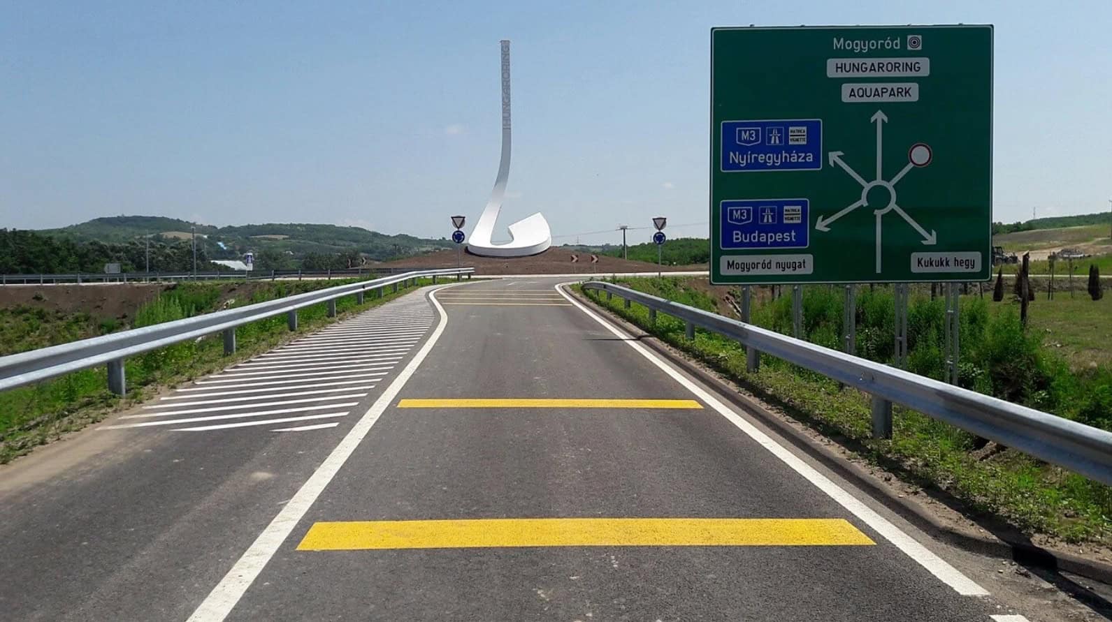 A road leading to the Hungaroring with directional signs for Mogyoród and Budapest, featuring a large, abstract monument in the center.