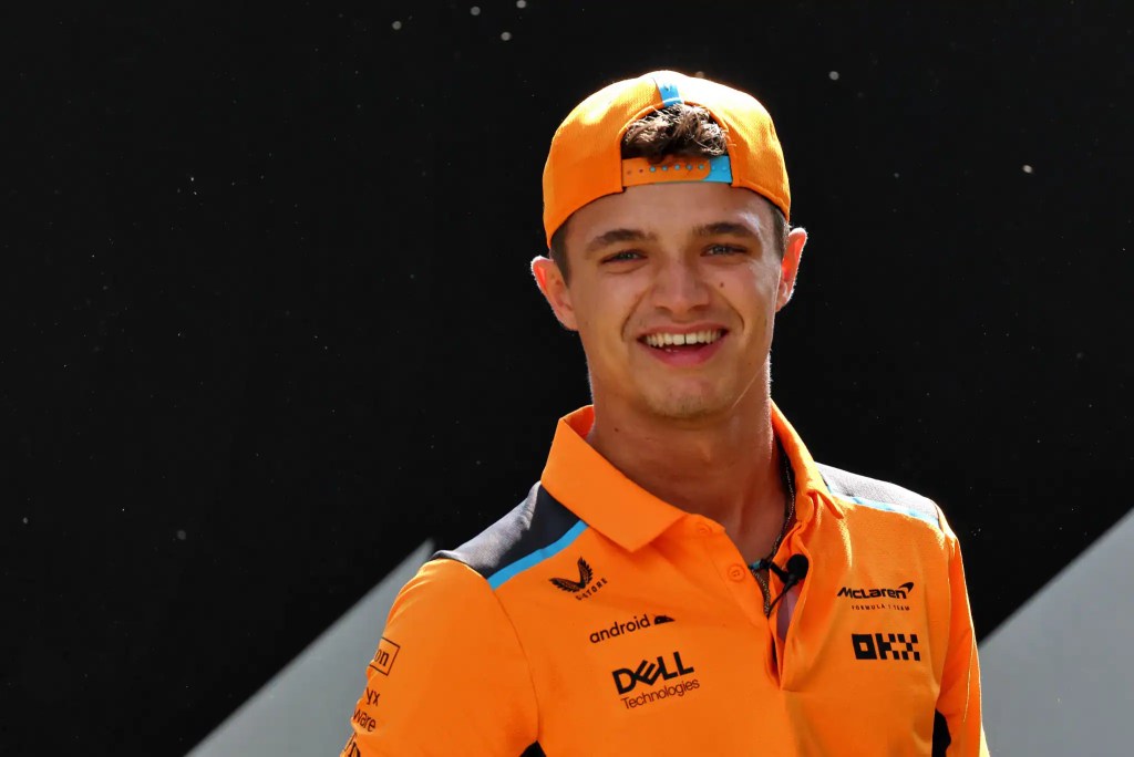 Lando Norris smiling in a McLaren racing outfit, showcasing his signature cap during a Formula 1 event.