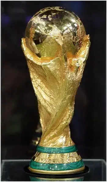 FIFA World Cup trophy displayed on a pedestal, featuring a golden globe held by two stylized hands, with green and gold accents at the base.
