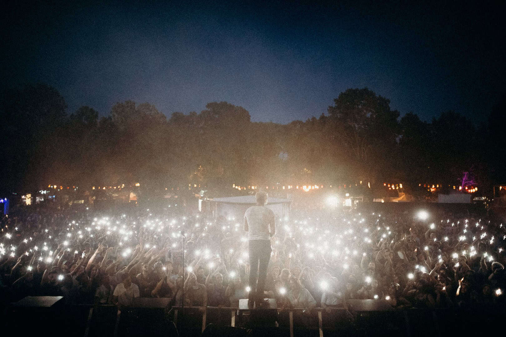 Large outdoor concert crowd at night with thousands of phone lights illuminated, forest backdrop, fe