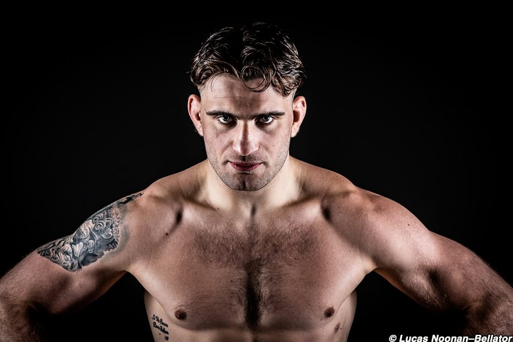 Man with tattoos looking intense, shirtless against a black background.