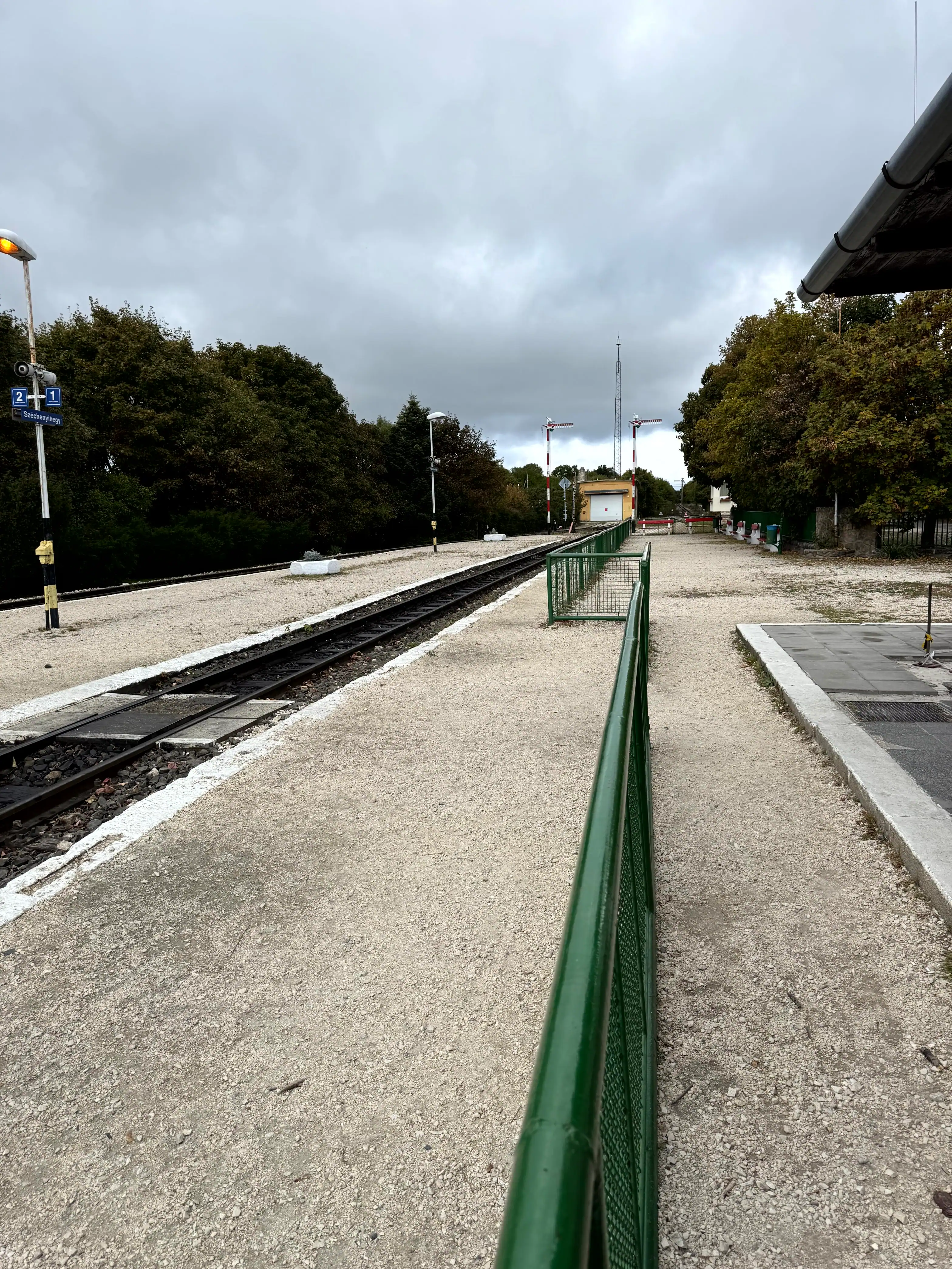Railway platform at Budapest airport station with trees and cloudy sky.