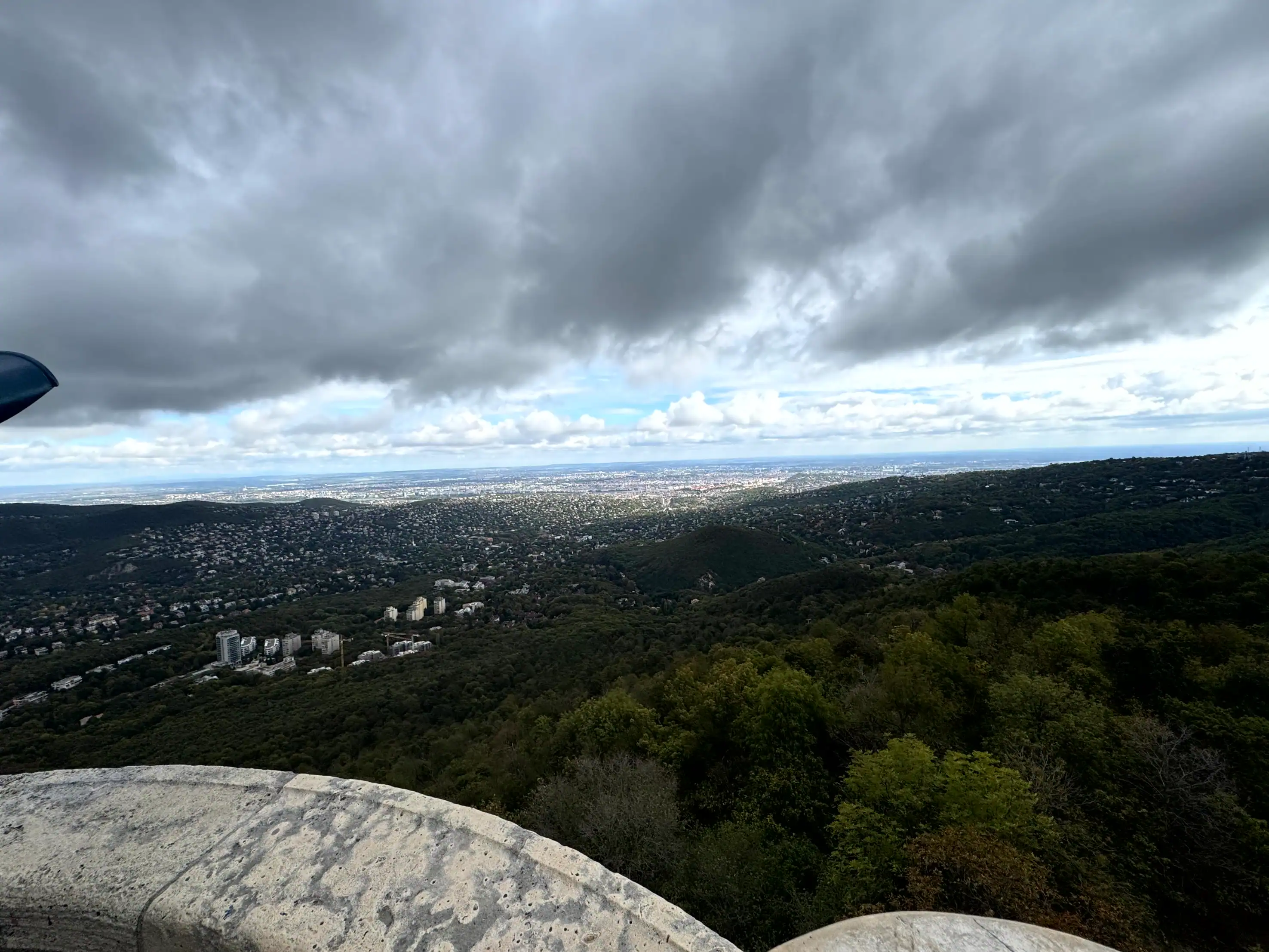 Scenic view from a hilltop overlooking Budapest with cloudy skies and lush greenery in the foregroun.