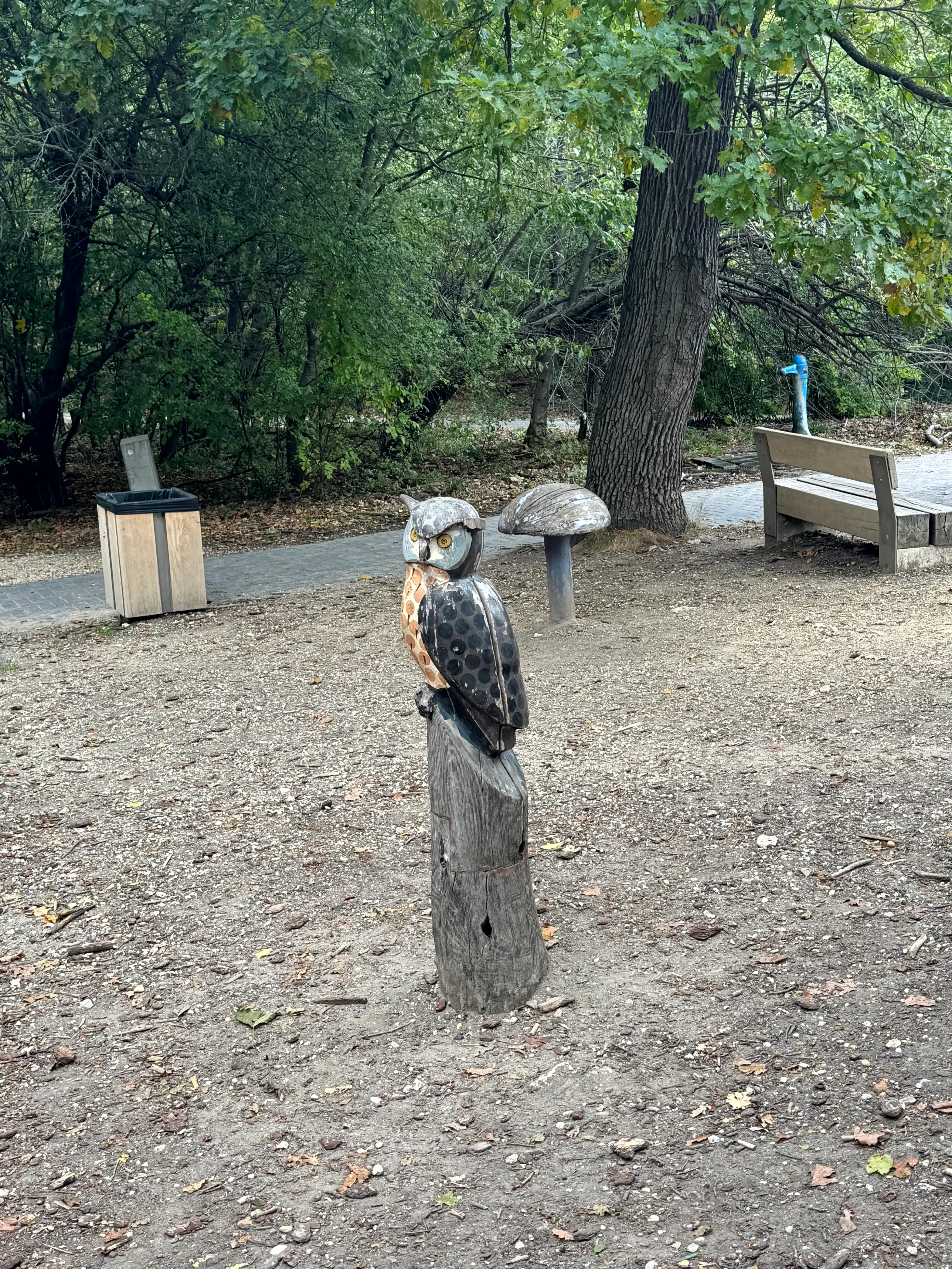 Owl sculpture perched on a wooden post in a park with trees and benches in the background.
