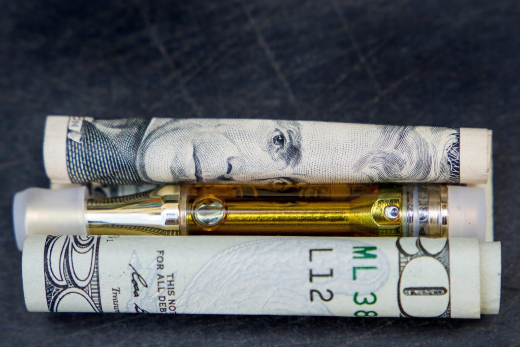 $100 bill, vape pen, and rolled-up dollar bills on dark surface - vaping, vape deals, and financial savings.