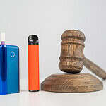 Vape device and e-cigarette standing next to a wooden gavel, symbolizing vaping regulations and legal considerations in the vaping community.