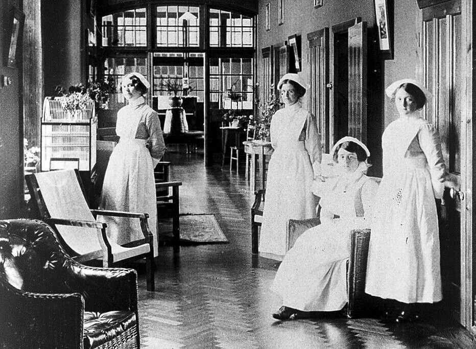Black and white photo of four nurses in vintage hospital uniforms inside a historic medical facility.