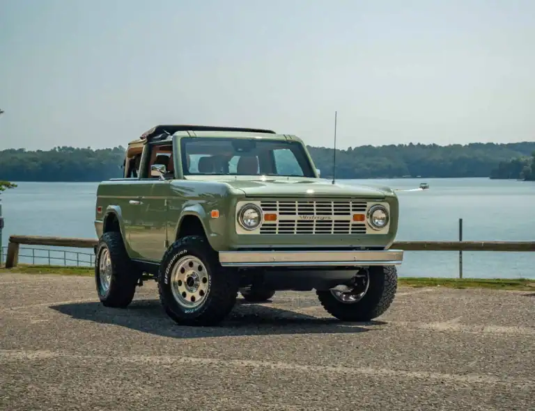 Vintage green off-road vehicle by a lakeside with a scenic water and forest background.