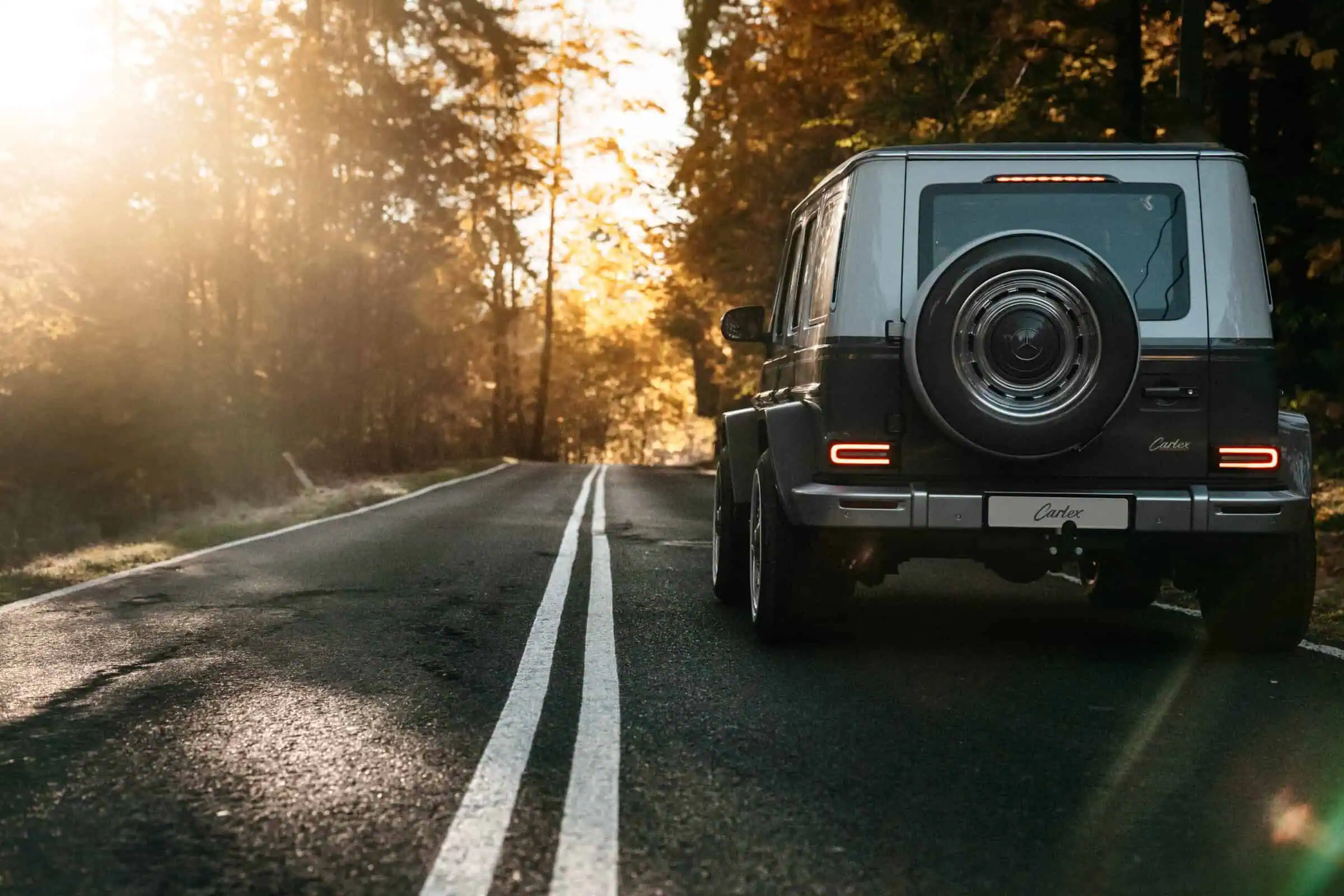 Luxury black Mercedes-Benz G-Class SUV parked on a scenic autumn road with golden sunlight filtering through trees, showcasing timeless automotive design and outdoor adventure appeal.