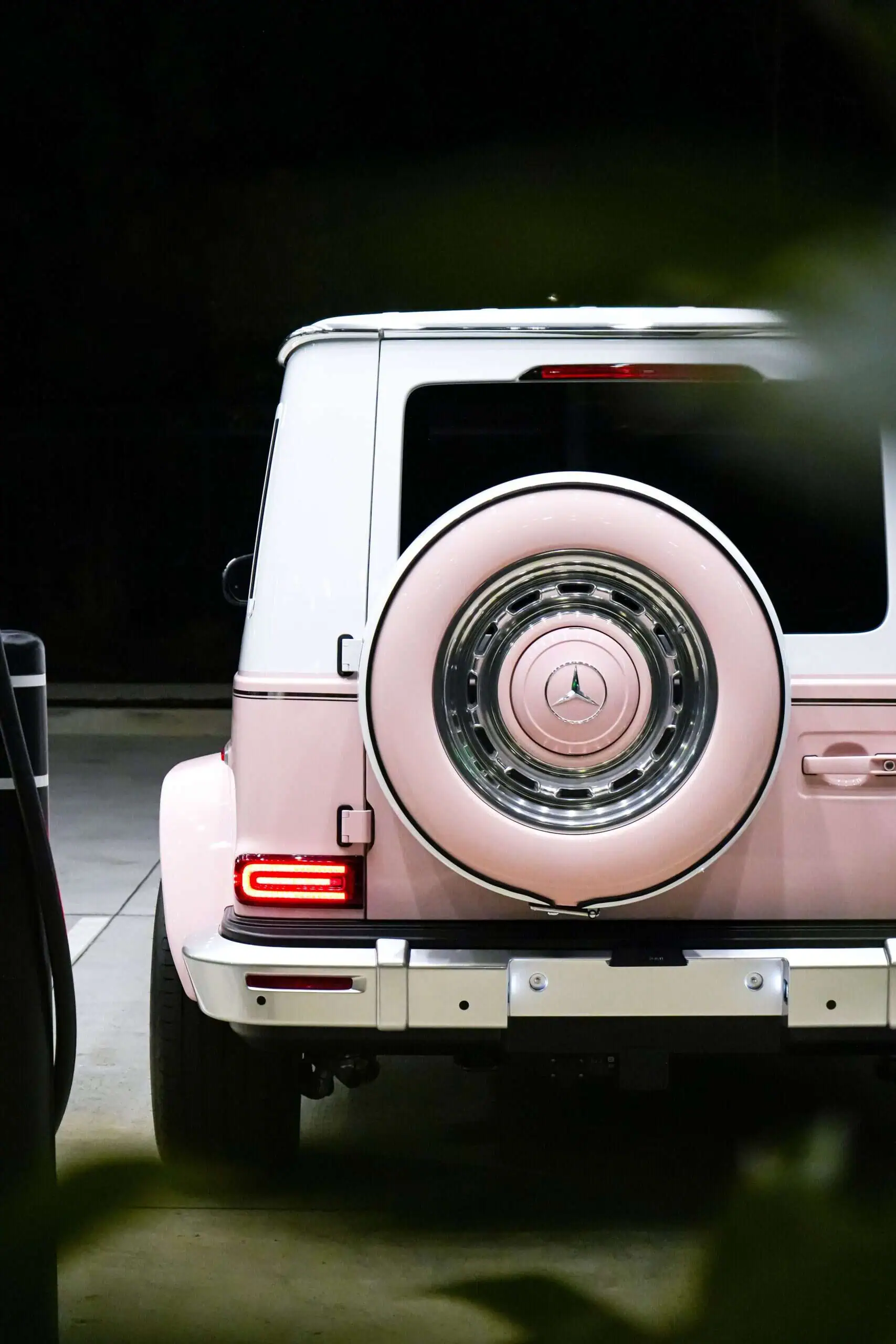 Pink luxury Mercedes-Benz G-Class SUV with custom design, rear view showcasing spare tire, nighttime urban setting, stylish and modern vehicle, perfect for luxury car enthusiasts.