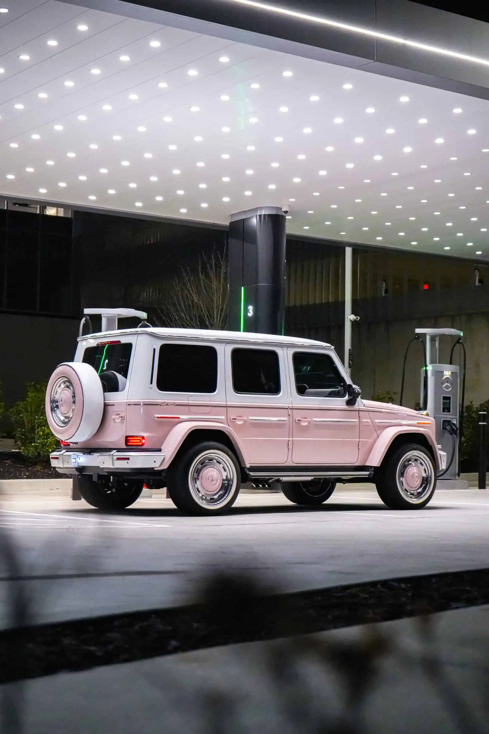 A pink Mercedes-Benz G-Class SUV, classic and modern design, parked at an electric vehicle charging station under a sleek, illuminated canopy showcasing vintage-modern automotive style.