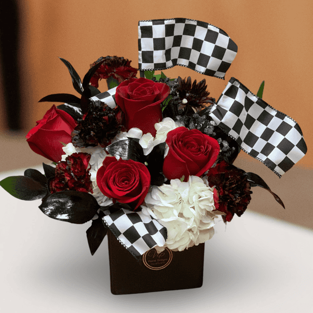 "Vegas Speedway Blossoms": A vibrant blend of red roses, black carnations, and white hydrangeas in a sleek black ceramic container adorned with checkered flags. The perfect Las Vegas Grand Prix centerpiece.