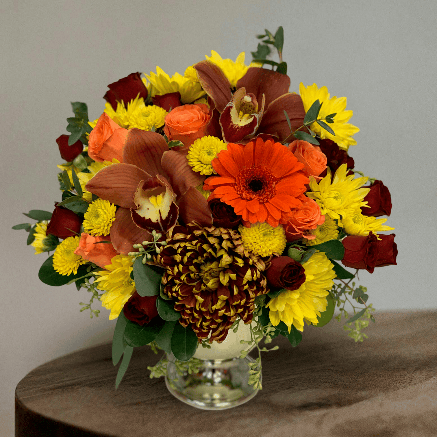 Vibrant fall floral arrangement with orange gerbera daisies, roses, orchids, chrysanthemums, and green foliage in a clear glass vase on wooden surface.