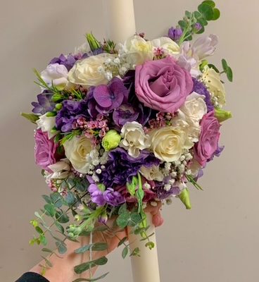 Luxury floral arrangement with purple, white, and pink roses, greenery, and a tall white candle, perfect for special occasions or romantic decor.