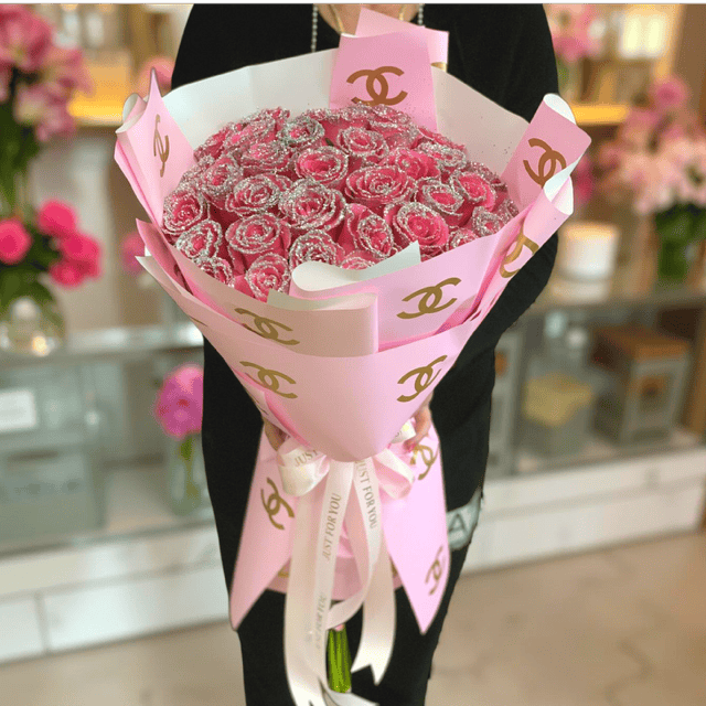 Pink Chanel-themed flower bouquet featuring a stunning arrangement of pink glitter roses, wrapped in elegant pink paper with gold Chanel logo accents, perfect for luxurious celebrations and special occasions.