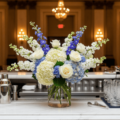 Elegant floral arrangement with white roses, hydrangeas, and blue delphiniums in a glass vase at a luxurious event venue, creating a sophisticated and inviting atmosphere.