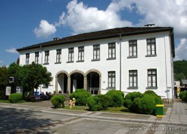 A spacious white building surrounded by lush greenery, featuring numerous trees and bushes.