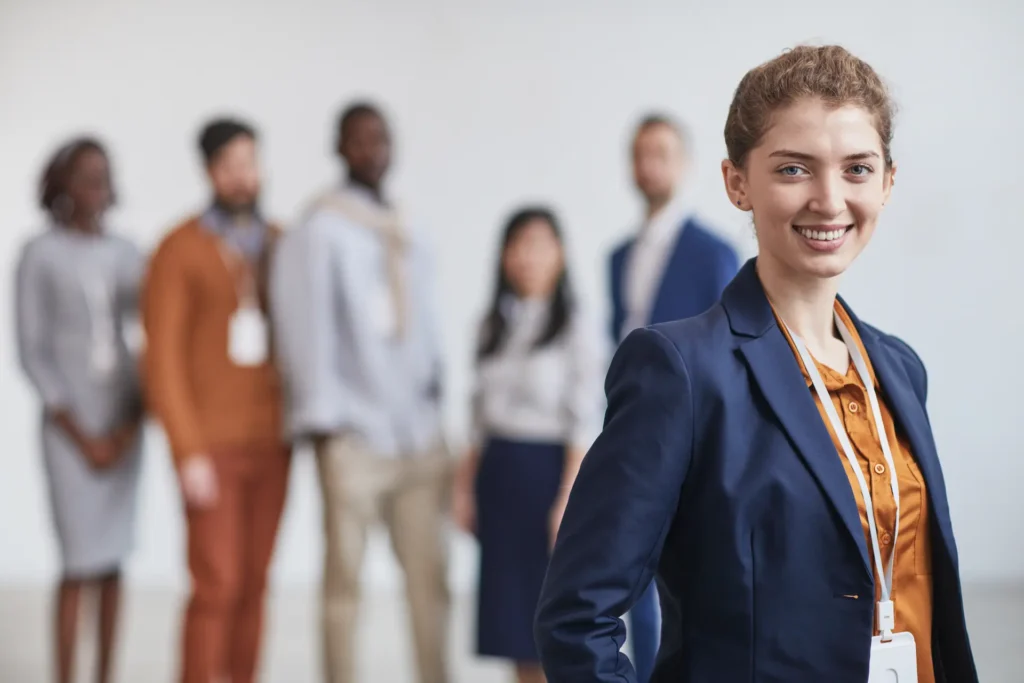 Professional young woman in navy business suit smiling confidently at camera with diverse group of professionals in background, representing career success and networking opportunities in recruitment apprenticeship programmes.