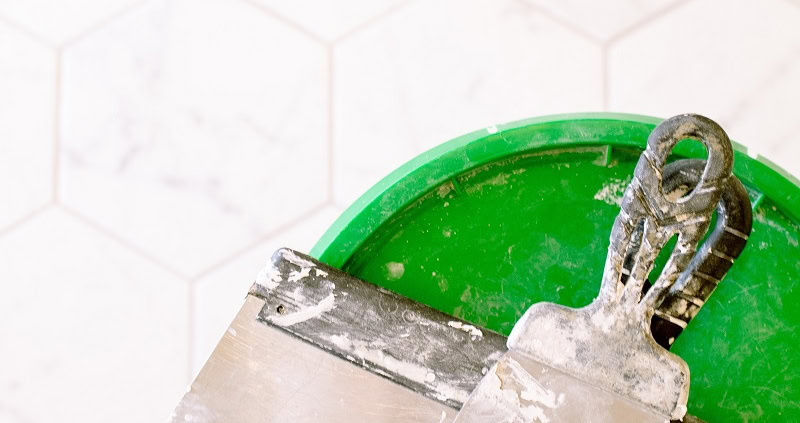 Tiling tools including a trowel and grout on a green bucket in a bathroom.