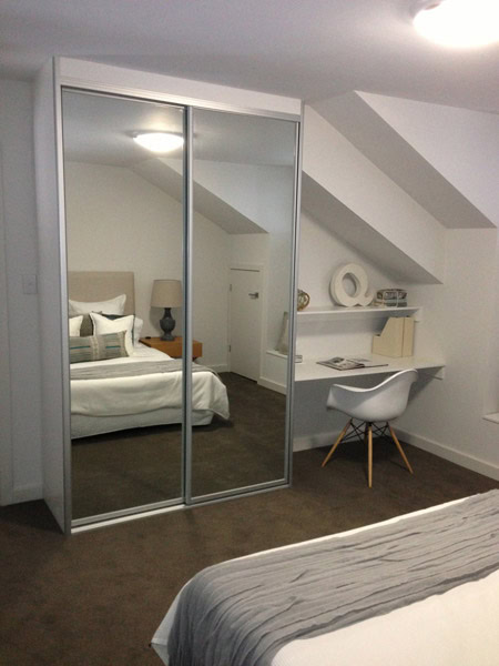Modern bedroom with built-in wardrobe featuring mirror sliding doors, a cozy bed, and a small worksp.