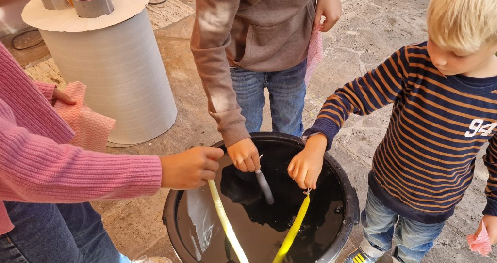 Farbige Wachskerzen beim Kerzenziehen, Kinder beim Kerzenmachen mit Wachs, kreative Bastelaktivität mit Wachs und Dochten, kindgerechtes Wachs-Workshop, nachhaltiges Kerzenhandwerk für Kinder.