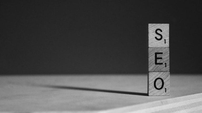 Black and white photo of three wooden Scrabble tiles stacked vertically to spell out the acronym "SEO".