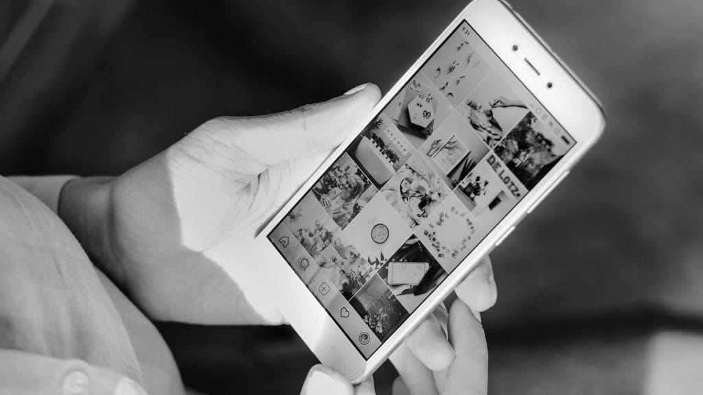 Black and white photo of a person's hands holding a smartphone and scrolling through an Instagram profile feed.