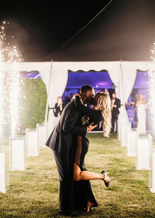 A couple shares a romantic dance at a luxury wedding reception under a beautifully decorated tent with celebratory fireworks.