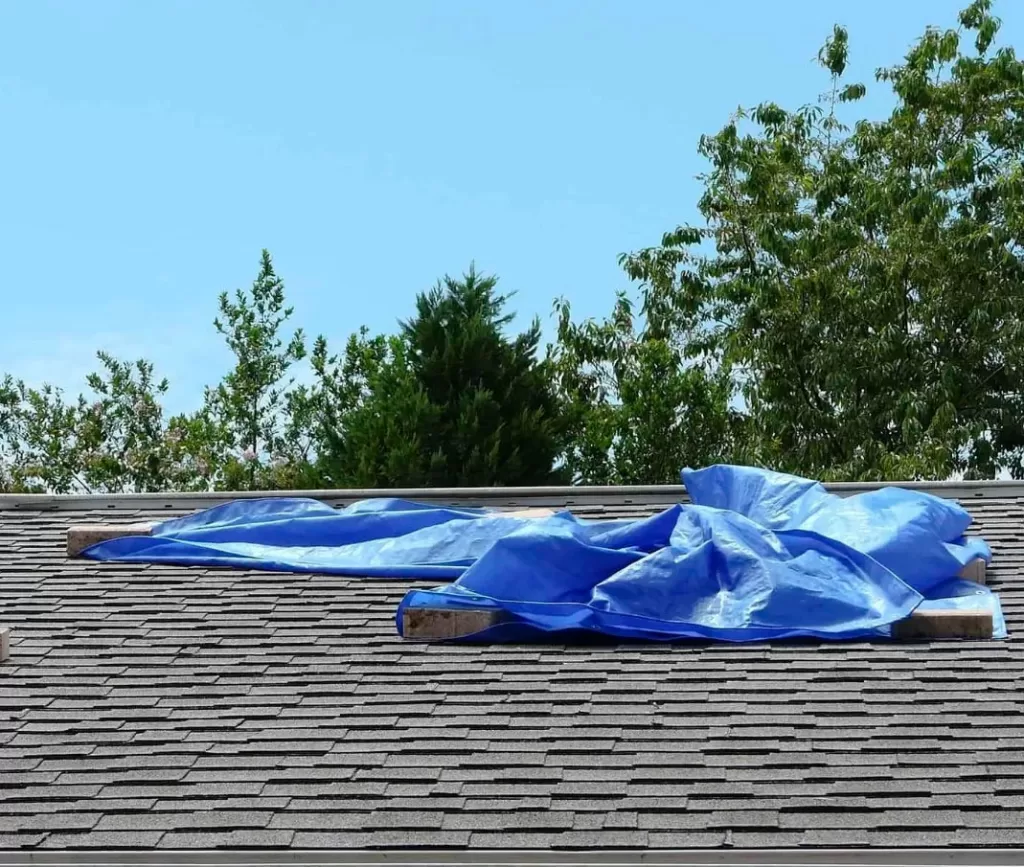 Storm Damage 6 A blue tarp held down by wooden planks covers part of a shingled roof, a clear sign of emergency storm damage repair, with trees and sky in the background.