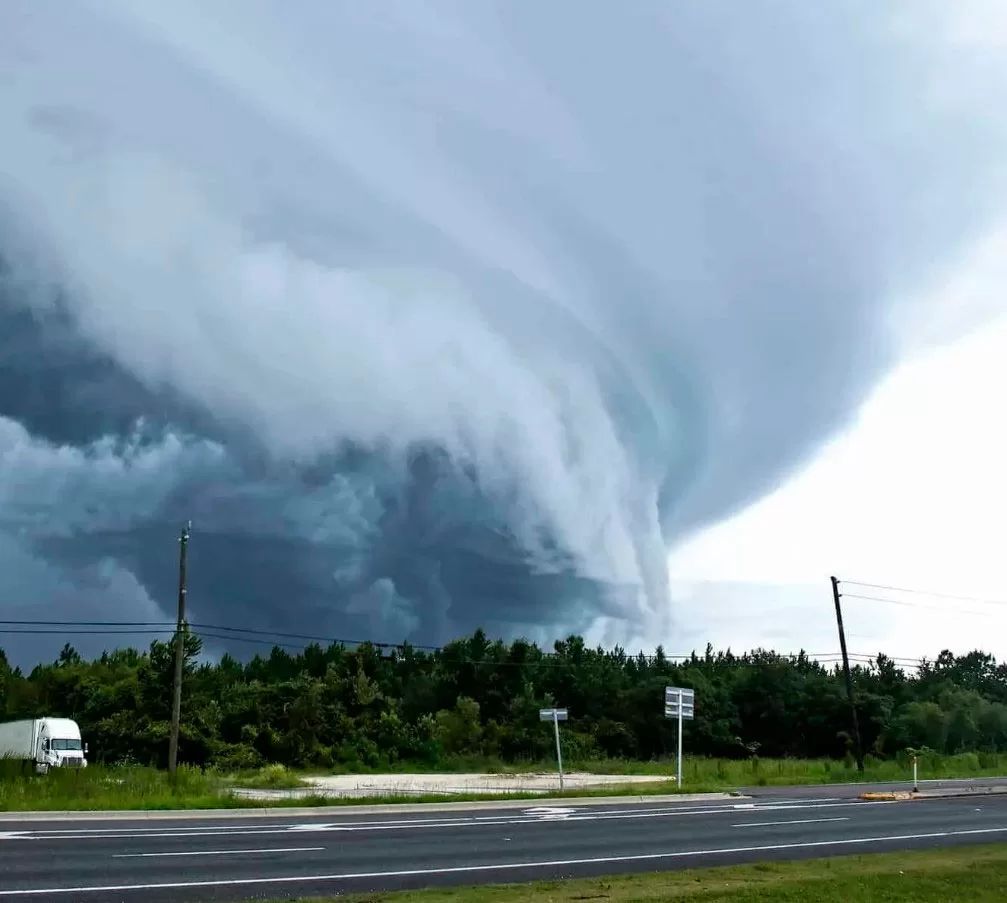 Storm Damage 7 A massive, swirling storm cloud formation looms over a green forest and a highway, with a white truck and road signs in the foreground—highlighting the urgent need for emergency storm damage repair after severe weather events.