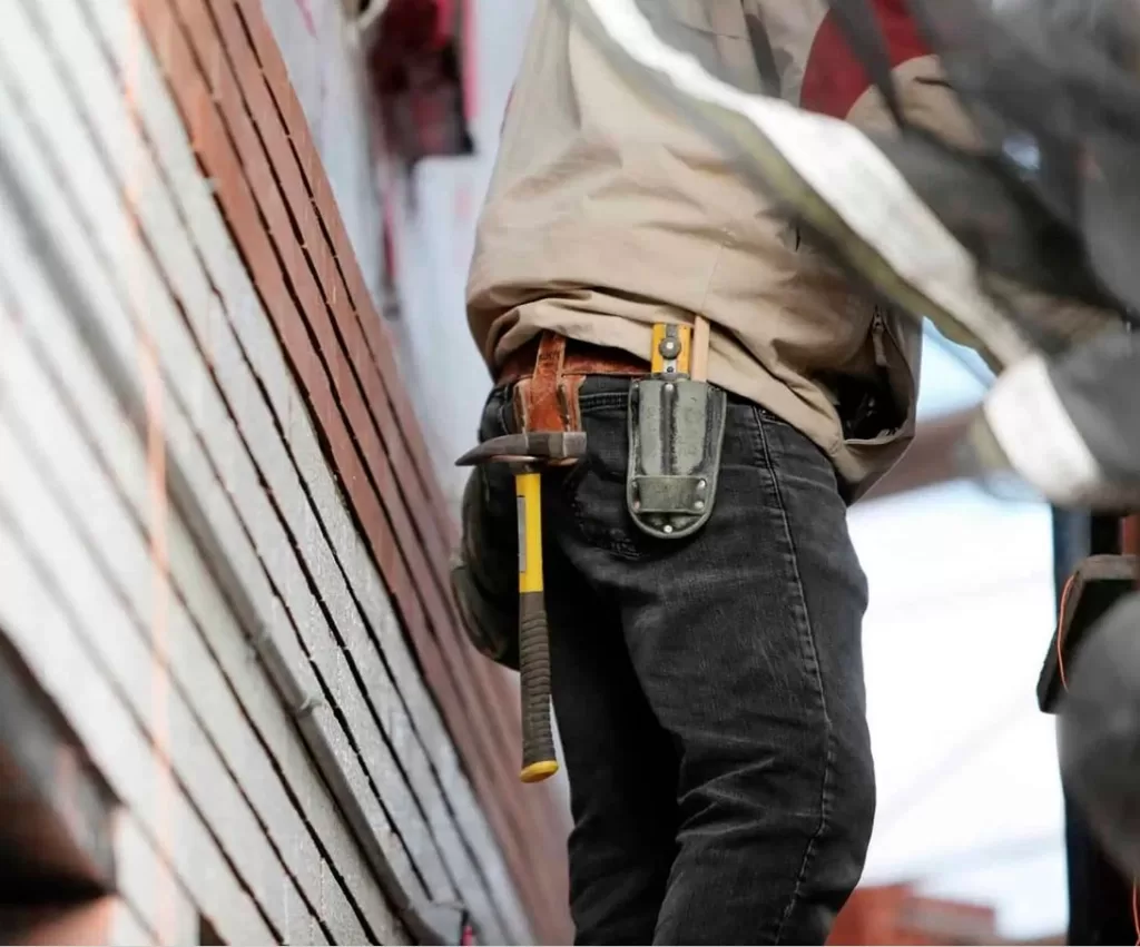 Storm Damage 11 Person in work clothes stands beside stacked bricks, with a hammer and tool pouch attached to their belt, ready for roof repair after storm damage.