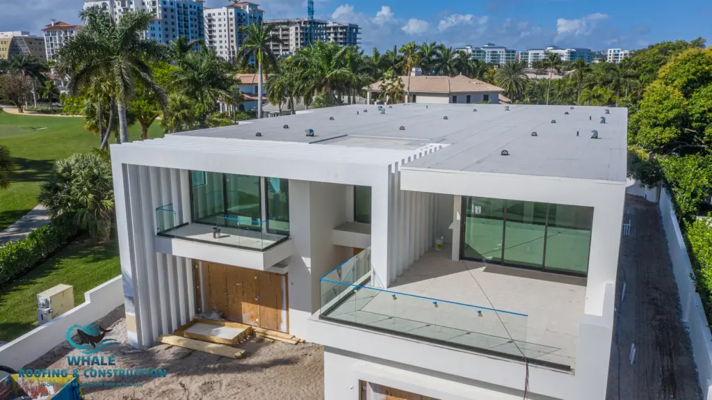 Flat Roof 4 A modern, two-story white building with large glass balconies and a flat roof is shown under construction, surrounded by palm trees and nearby high-rise buildings—ideal for those seeking expert flat roof repair.