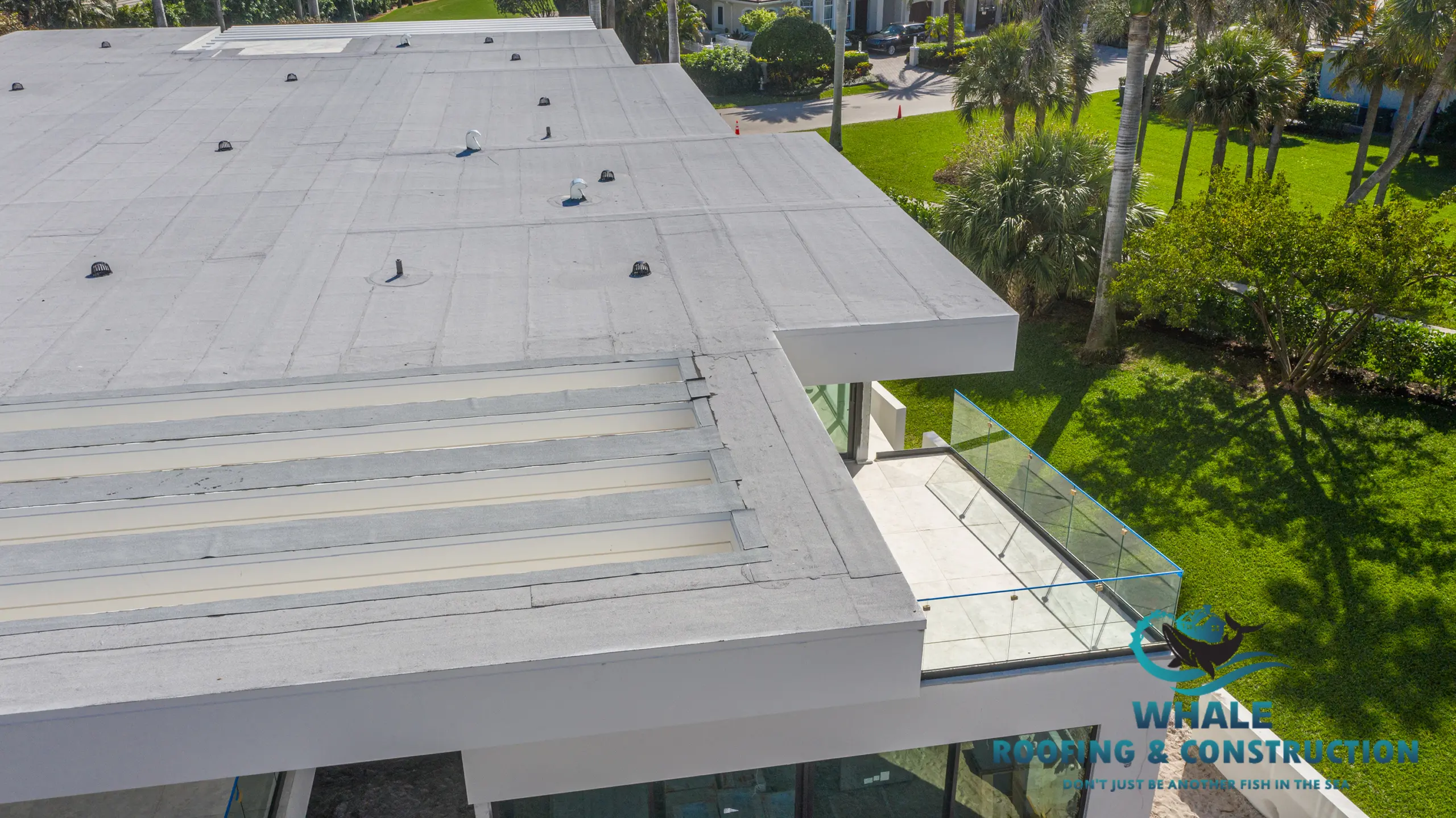 Commercial Roofing Services in Boca Raton, FL 6 Aerial view of a flat commercial building roof with skylights, pipes, and a glass balcony—great for assessing flat roof replacement cost—surrounded by grass and trees.