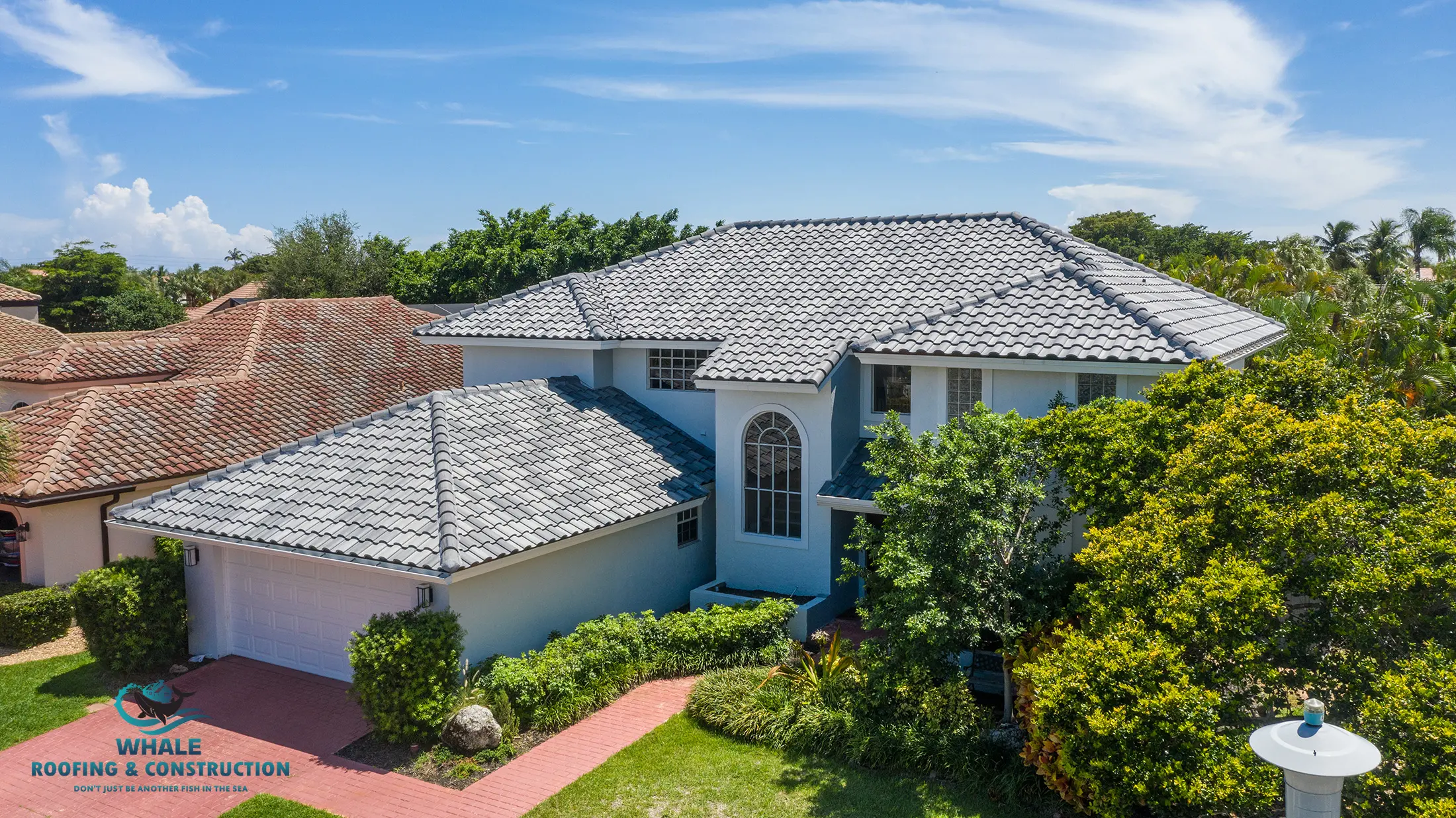 A two-story light blue house with grey tiled roof, surrounded by trees and shrubs, with a red brick driveway and a Whale Roofing & Construction logo in the corner.