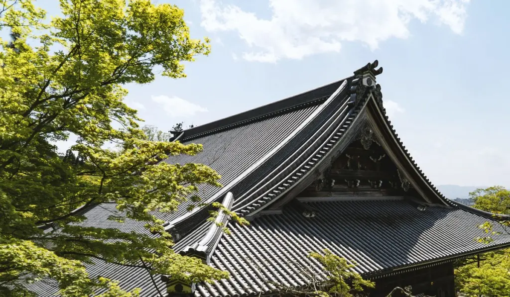 Storm Damage 2 Traditional Japanese temple roof with ornate details, surrounded by green leafy trees under a partly cloudy sky.