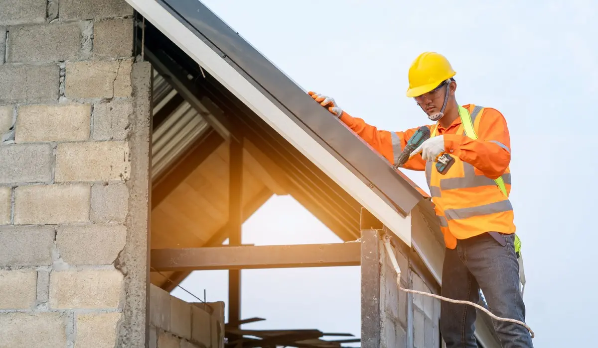 A construction worker in a safety vest and helmet inspects or measures the edge of a rooftop on a partially built structure.