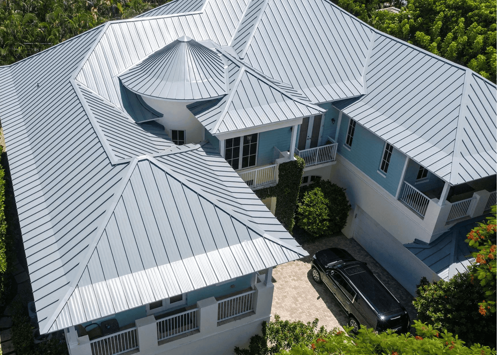 Roof Repair 2 Aerial view of a large blue house with a complex metal roof design and a black car in the driveway, surrounded by greenery—showcasing expert roof maintenance Boca Raton for lasting beauty and protection.