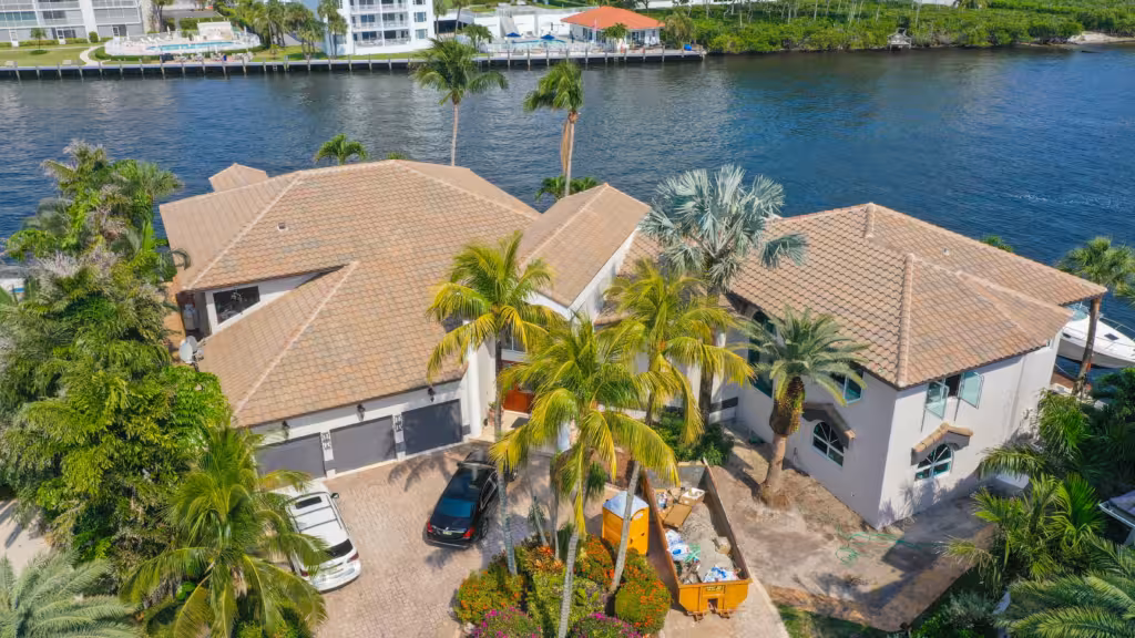 Aerial view of a large waterfront house with a tile roof, multiple palm trees, parked cars, and a dumpster in the driveway, next to a canal.