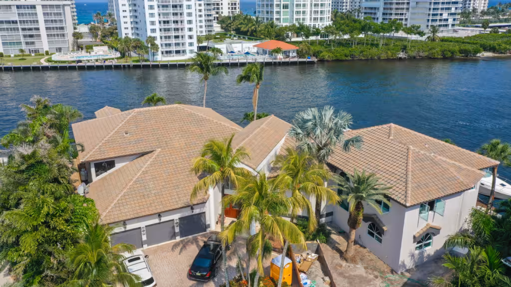 Tile Roofing 2 Aerial view of a waterfront house with stunning tile roofing, surrounded by palm trees near a canal, with high-rise buildings in the background. Perfect inspiration for your next tile roof replacement or repair project.