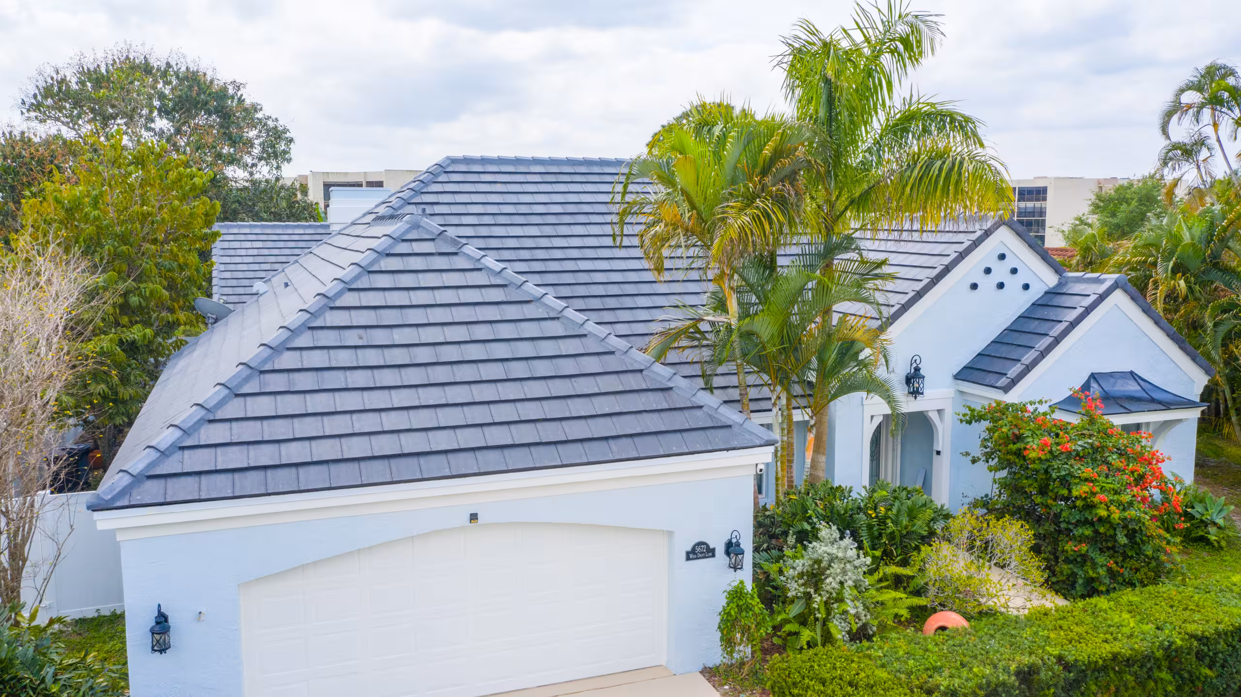 A light blue house with a dark gray tiled roof, white garage door, and lush green landscaping with palm trees and bushes in front.