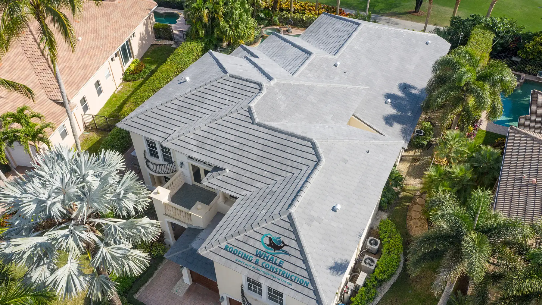 Roof Financing 7 Aerial view of a large house with a gray shingle roof featuring multiple angles and ridges, surrounded by palm trees and neighboring homes.