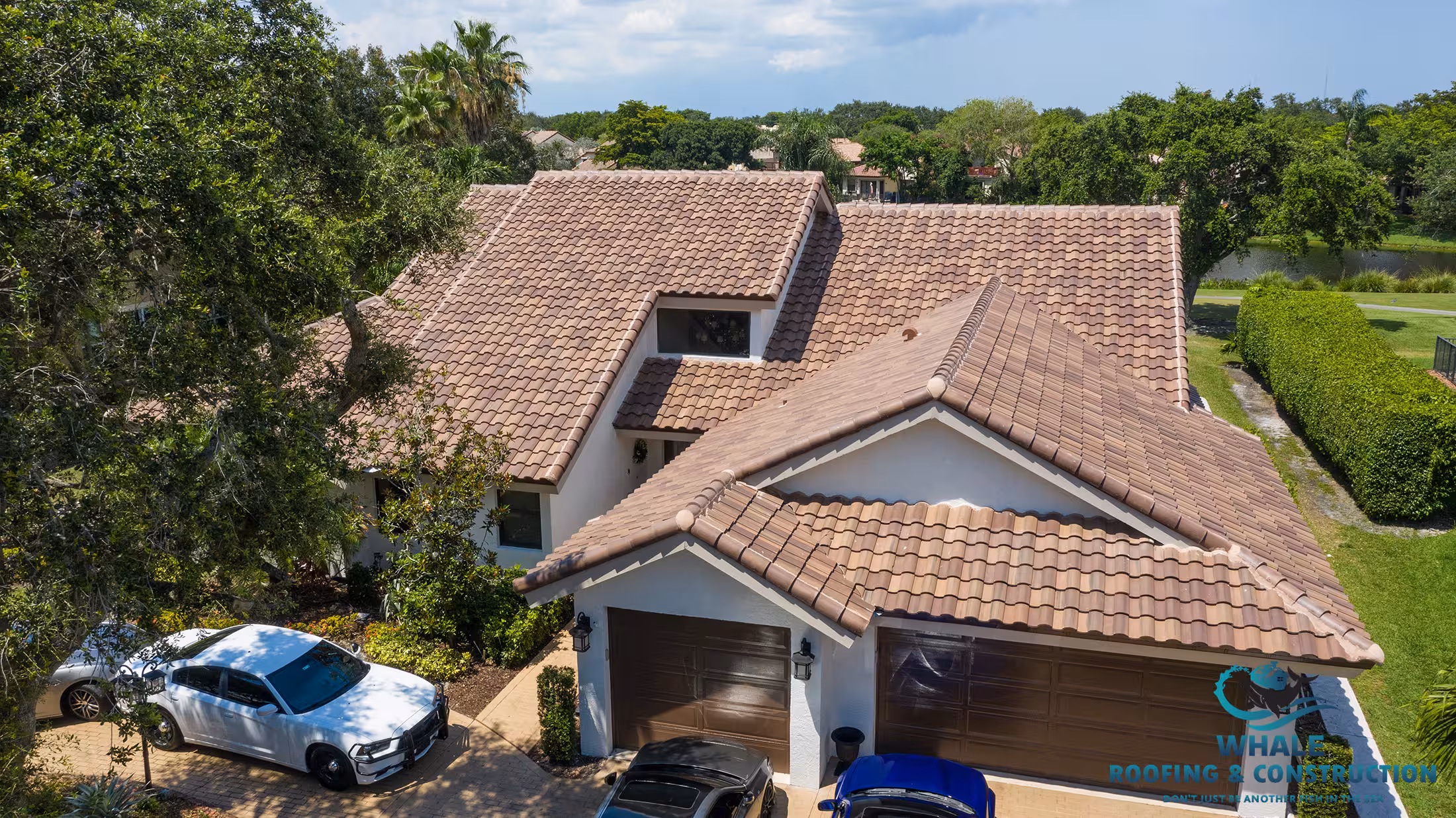 Aerial view of a house with a brown tile roof, attached garage, and several cars in the driveway; trees and a lake are visible in the background—ideal for those seeking tile roofing services or expert tile roof installation.