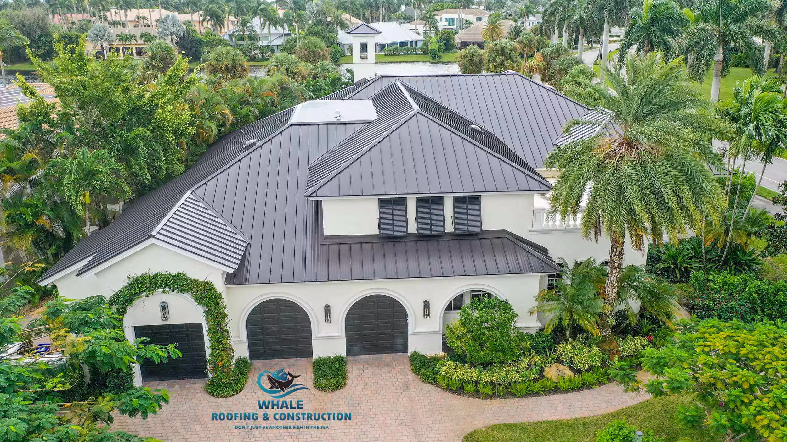 A large, two-story house with a dark metal roof, white exterior, three black garage doors, and surrounded by lush green trees and landscaping.