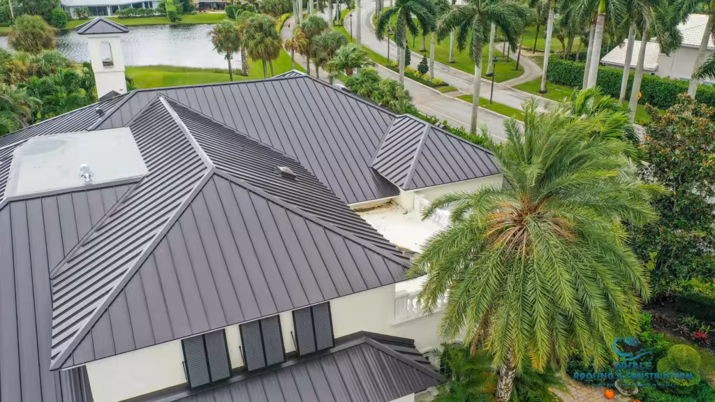 Aerial view of a modern house with a dark metal roof surrounded by palm trees and landscaping near a road and water.