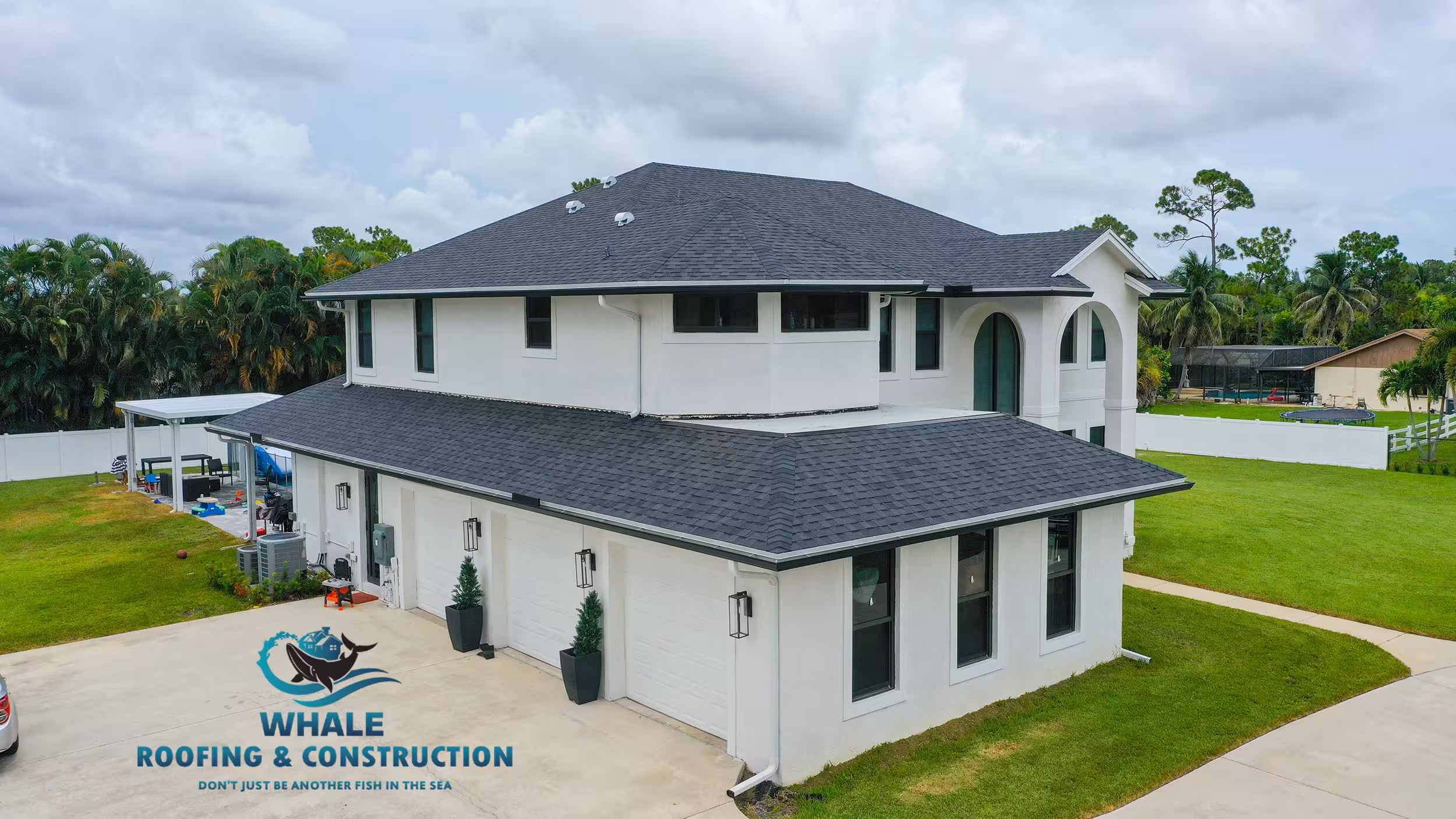 A two-story white house with dark gray shingle roofing sits on a well-manicured lawn; Whale Roofing & Construction logo appears in the left corner, highlighting the best shingle roofing contractors in the area.